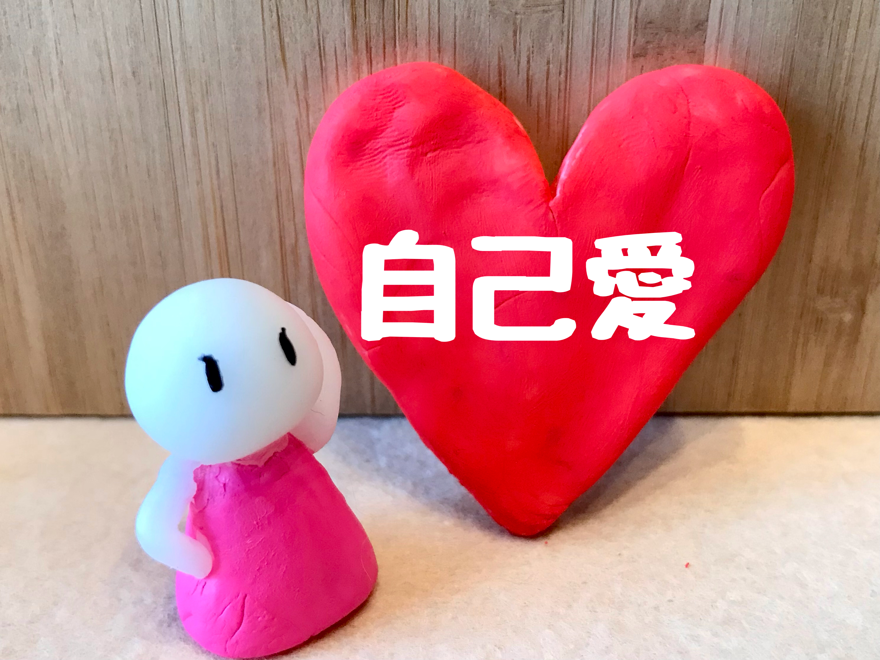 「自己愛」と白文字で書かれた大きな赤い粘土のハートの前に、誇らしげな表情のクレイモデルが立っている画像